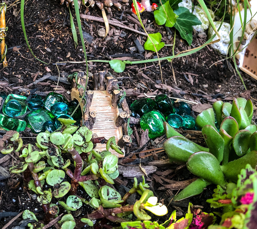 Outdoor fairy garden with a miniature bridge and blue gemstones to mimic water.